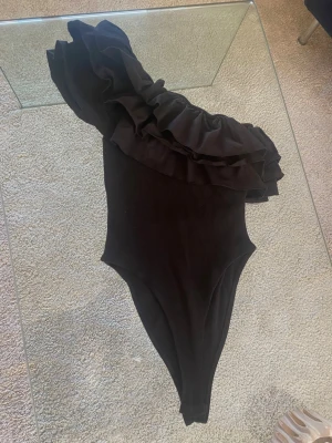Svart asymmetrisk body med volang - ACCEPTERAR BUD PÅ 400kr!Säljer en svart body med en axel och stora volangdetaljer längs urringningen. Bodyn har en tight passform och är tillverkad i stretchigt material. Perfekt för dig som vill ha något unikt och trendigt i garderoben. Använd 1 gång.