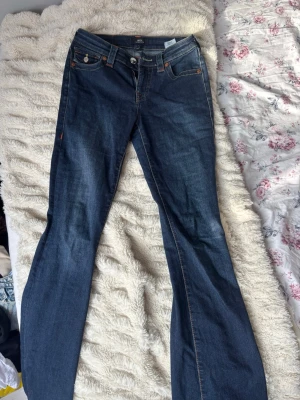 Blå bootcut jeans från True Religion - Snygga blå bootcut jeans från True Religion med klassiska kontrastsömmar och dekorativ ficka bak. Jeansen har normal passform och är tillverkade i bomull med en mörk tvätt. Modellen heter BECCA Mid Rise Bootcut 