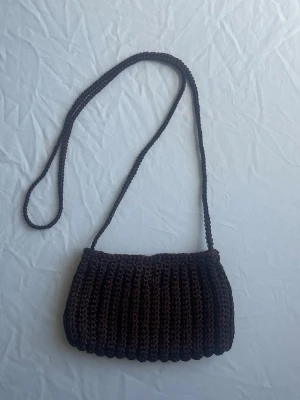 Brun virkad axelväska - Snygg brun axelväska i virkat material med lång axelrem. Väskan har en mjuk, ribbad struktur och stängs med magnetlås. Perfekt för dig som gillar en unik och handgjord look.