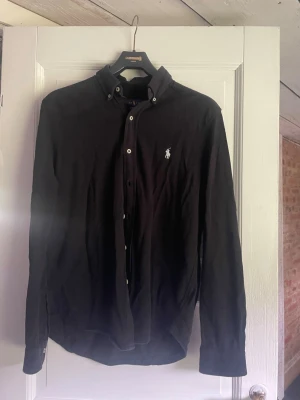 Svart skjorta från Ralph Lauren linne  - Klassisk svart skjorta från Ralph Lauren med button-down krage och vit broderad logga på bröstet. Skjortan har långa ärmar och knäppning framtill. Perfekt till en stilren och enkel look.