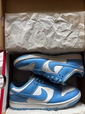 Nike Dunk Low UNC blå/vit - Nike Dunk Low sneakers i färgen UNC blå och vit. Skorna har klassisk låg siluett, vit tåbox med perforeringar och blått läder runtom. Swoosh-logga på sidan och matchande blå snörning. Yttersula i gummi och ovandel i läder för en clean och sportig look.