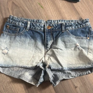 Blå denimshorts med slitningar - Säljer ett par snygga blå denimshorts med slitningar och fransig kant. Shortsen har fem fickor, knappgylf och en cool tvättad look som ger en avslappnad vibe. Perfekta för sommardagar och festivaler! Midjebandet i har blivit slitet men går lätt att byta ut