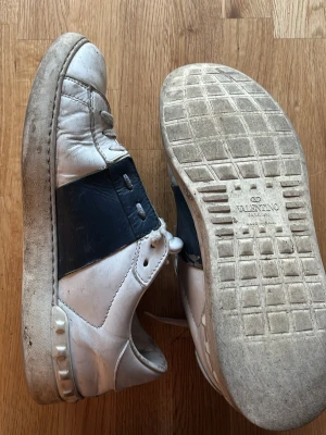 Valentino vita och svarta sneakers - Snygga sneakers från Valentino i vitt läder med en bred svart läderdetalj över sidan. Klassisk låg modell med rund tå och vita snören. Sulan är vit med diskreta nitar på hälen. Märkeslogga på plösen och yttersulan. Tar emot BYTEN!