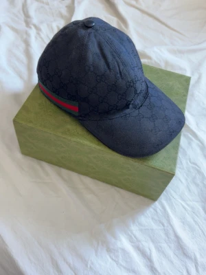Svart Gucci keps med GG-mönster - Snygg svart keps från Gucci med det klassiska GG-monogrammet över hela tyget. Kepsen har en böjd skärm och en grön-röd rand på sidan som ger en ikonisk touch. Tillverkad i bomullscanvas och har justerbar rem bak. Har tvättat den förut så tyget i skärmen är lite löst plus att kepsen är lite smutsig. Har orderbekräftelse kom privat om du vill se eller har några andra frågor.👍