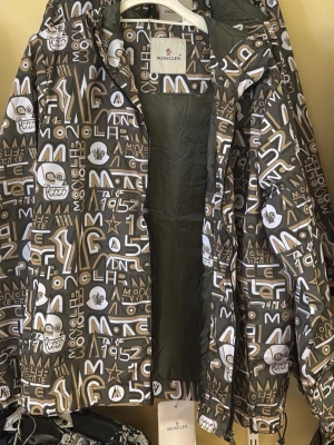 Moncler grafisk vindjacka med huva - Säljer en unik vindjacka från Moncler med grafiskt mönster i brunt, vitt och beige. Jackan har huva, dragkedja och coola tryck med loggor och figurer. Materialet är lätt och vattenavvisande, perfekt för blåsiga dagar. Insidan har en serietidningsetikett som signaturdetalj.