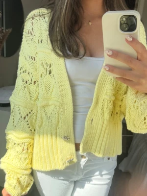 Gul stickad kofta med mönster - Söt ljusgul stickad kofta med hålmönster och dekorativa knappar framtill. Koftan har ballongärmar och ribbade muddar, samt en croppad passform. Perfekt för att liva upp din outfit med en färgklick. Materialet känns mjukt och luftigt. Saknar knappen högst upp men passar super trotts det