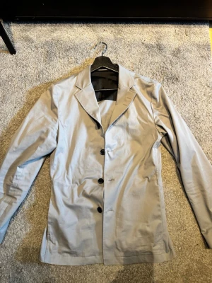 Tiger of Sweden overshirt/kavaj - Stilren ljusgrå kavaj/ overshirt med tre svarta knappar framtill och klassisk krage. Kavajen har lång ärm och är tillverkad i ett slätt tyg med lätt glans. Passar perfekt till en dressad look och har en enkel, tidlös design.