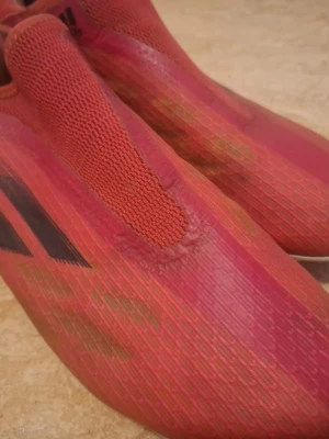 Adidas röda fotbollsskor X-serien - Säljer ett par röda Adidas fotbollsskor från X-serien med strumpliknande passform och låg profil. Skorna har en slimmad design utan snörning, svart Adidas-logga på sidan och ovandel i syntetmaterial för bra bollkontroll. Perfekta för snabba spelare som vill ha lättviktsskor.