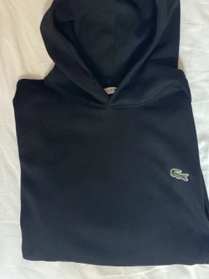 Svart hoodie från Lacoste - Snygg svart hoodie från Lacoste med klassisk krokodillogga på bröstet. Regular fit-modell med huva och mjukt material som känns skönt mot huden. Perfekt för dig som gillar stilrena och tidlösa plagg med en sportig touch.