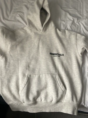 Grå Essentials Fear of God hoodie - Snygg ljusgrå hoodie från Essentials Fear of God med svart logga på bröstet och stor text på ryggen. Klassisk känguruficka framtill och stor huva. Tillverkad i mjukt bomullsmaterial som känns riktigt skönt mot huden. Perfekt för en avslappnad streetwear-look.
