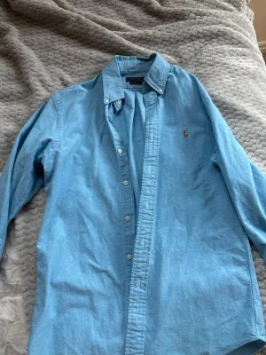 Blå oxfordskjorta från Polo Ralph Lauren - Säljer en klassisk ljusblå oxfordskjorta från Polo Ralph Lauren i slim fit. Skjortan har knappar hela vägen, button-down krage och den ikoniska broderade loggan på bröstet. Tillverkad i bomull med stretch för extra komfort.