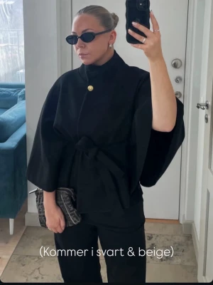 Svart cape-kappa med bälte - Har aldrig använt nästan, har lite annorlunda stil nu. Kappan har vida ärmar och hög krage, vilket ger en elegant och modern look. Perfekt för dig som vill ha en trendig och unik ytterjacka.