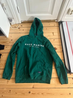 Grön hoodie från Race Marine - Snygg grön hoodie från Race Marine med vit logga på bröstet och känguruficka framtill. Klassisk huva med dragsko och ribbade muddar vid ärmar och nederkant. Tillverkad i mjuk bomullsblandning, perfekt för en avslappnad stil.