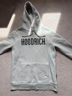 Ljusgrå hoodie från Hoodrich - Snygg ljusgrå hoodie från Hoodrich med stor svart logga på bröstet. Hoodien har en klassisk huva med dragsko och en stor magficka framtill. Tillverkad i mjukt material som känns skönt mot huden. Perfekt för en avslappnad streetwear-look.
