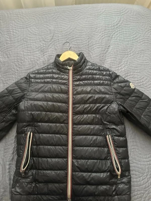 Mörkblå dunjacka från Moncler (äkta) - Snygg navy blue dunjacka från Moncler med quiltad design och ståkrage. Jackan har dragkedjor med röd, vit och blå detalj samt Moncler-logga på ärmen. Tillverkad i lätt och värmande material, perfekt för våren, kalla sommarkvällar och hösten. Storlek är medium/slim fit.