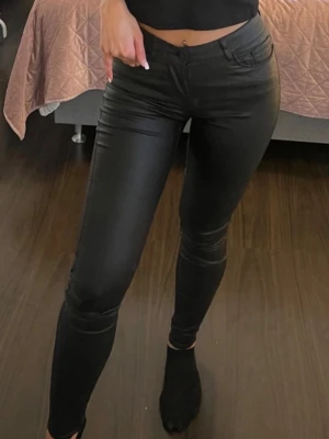 Svarta skinny jeans i coated material - Säljer ett par svarta skinny jeans med coated finish som ger en cool läderlook. Skinnimitation, Jeansen har hög midja, fem fickor och smal passform hela vägen ner. Perfekta för dig som vill ha en trendig och edgy stil.storlek S