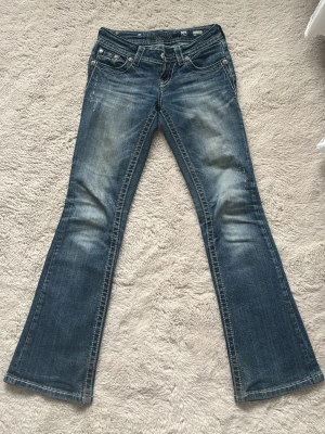 Blå bootcut jeans med broderi - Säljer ett par blå bootcut jeans med snygg slitning och broderade detaljer på bakfickorna. Jeansen har klassisk femficksmodell och är tillverkade i denim med stretch. Perfekta för dig som gillar en avslappnad men trendig look.