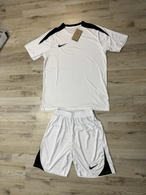Vitt Nike träningsset med svarta detaljer - Säljer ett fräscht vitt träningsset från Nike med svarta detaljer på axlar och shorts. Setet består av en kortärmad t-shirt och matchande shorts i lätt och snabbtorkande material, perfekt för fotboll eller gym. Klassisk Nike-logga på både tröja och shorts.