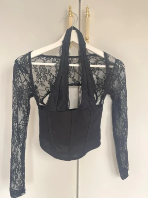 Svart spetsblus med djup urringning - Trendig svart blus med långärmade spetsärmar och djup urringning Kroppsdelen är i slät, figurnära tyg medan ärmar och axelparti är i transparent spets med blommönster. storlek: XXS men är mer som XS. Aldrig använd