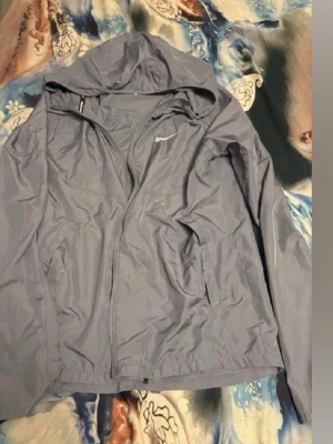 Ljuslila vindjacka från Nike - Snygg ljuslila vindjacka från Nike med huva och dragkedja framtill. Jackan har två sidofickor med dragkedja och en liten vit Nike-logga på bröstet. Tillverkad i lätt och tunt material som passar perfekt för blåsiga dagar.