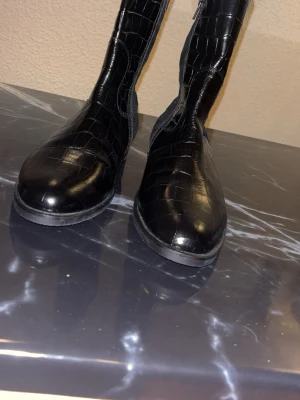 Svarta boots i krokopräglat skinn Gabor - Snygga svarta stövlar från Gabor i krokopräglat skinn. Modellen har rund tå, låg klack och dragkedja på sidan för enkel påtagning. Perfekta för dig som vill ha en trendig och stilren look med lite extra edge.