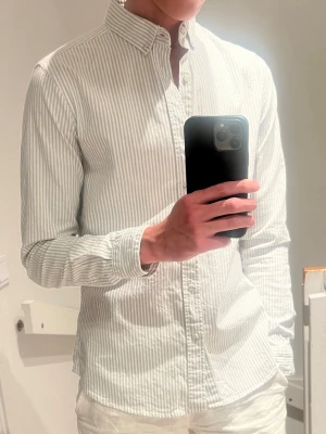 Vit och grå randig skjorta - Säljer en vit skjorta med tunna grå ränder och klassisk krage. Skjortan har lång ärm, knappar framtill och är tillverkad i mjuk bomull. Perfekt för en clean och fräsch look.