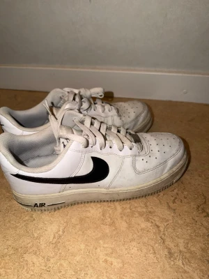 Nike Air Force 1 vita sneakers - Klassiska vita Nike Air Force 1 sneakers med svart swoosh och AIR-detalj på sulan. Skorna har lågt skaft, perforerad tåbox och är tillverkade i läder med gummisula. Perfekta för en clean och tidlös streetstil.