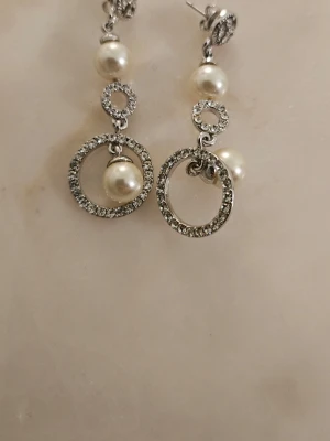 Silverfärgade örhängen med pärlor - Snygga örhängen i silverfärg med gnistrande stenar och vita pärlor. Hängande design med cirkelformade detaljer och pärlor som ger en elegant look. Perfekta för att lyfta din outfit med lite extra bling. Har aldrig användts.  Har allergi.  