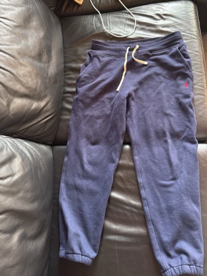 Blå mjukisbyxor från Polo Ralph Lauren - Mörkblå mjukisbyxor från Polo Ralph Lauren med dragsko i midjan och resår vid bensluten. Byxorna har två sidofickor och en liten röd logga broderad på vänster sida. Perfekta för chill dagar eller när du vill vara extra bekväm.