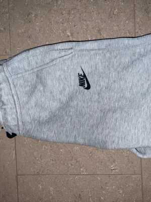 Grå mjukisbyxor från Nike - Snygga ljusgrå mjukisbyxor från Nike med klassisk logga på benet. Byxorna har resår i midjan och är tillverkade i mjukt bomullsmaterial som passar perfekt för chill eller träning. Enkel och stilren design som funkar till det mesta.