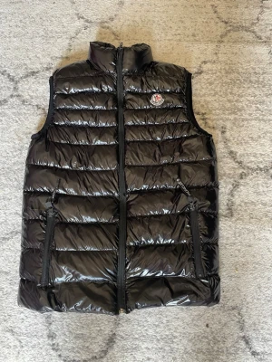 Svart Moncler väst - Snygg svart dunväst från Moncler med glansig finish och hög krage. Västen har två dragkedjefickor framtill och Moncler-logga på bröstet. Perfekt att slänga över en hoodie eller tröja för extra värme.