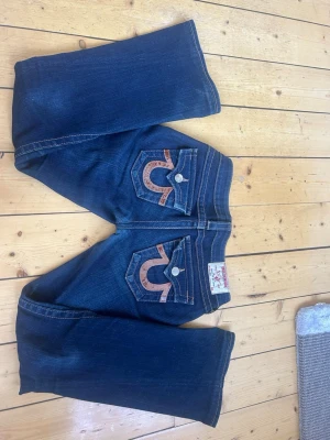 Mörkblå True Religion jeans - Säljer ett par mörkblå jeans från True Religion med ikoniska kontrastsömmar och stora broderade fickor bak. Klassisk femficksmodell med lite utsvängda ben och detaljerade knappar. Färgen har försvunnit lite från knäna efter användning men mycket fint skick annars