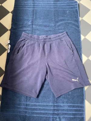 Mörkblå shorts från Puma med logga - Snygga mörkblå shorts från Puma med elastisk midja och två sidofickor. Klassisk Puma-logga i vitt på vänster ben. Perfekta för sommaren och träning, tillverkade i mjuk bomullsblandning för maximal komfort.