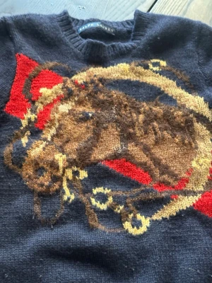 Mörkblå stickad tröja Ralph Lauren - Stickad tröja från Ralph Lauren i mörkblått med ett stort motiv av en häst och tyglar i brunt, beige och gult samt röda detaljer. Tröjan har rund halsringning och ribbstickade muddar vid ärmslut och nederkant. Perfekt för dig som gillar klassisk stil med en twist. Storlek 8-11 år ca 