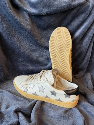 Saint laurent sneakers - Snygga vita sneakers från Saint Laurent med silverfärgade stjärnor på sidorna och svart detalj på hälen. Perfekta för sommaren, storlek 41,5 men passar bättre till 42
