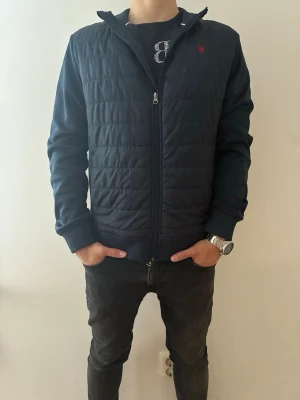 Ralph Lauren cardigan - Snygg mörkblå cardigan från Ralph Lauren med quiltad front och rygg, dragkedja hela vägen och diskret röd logga på bröstet. Jackan har ribbade muddar och är tillverkad i ett mjukt material som ger en sportig och stilren look. Minimal missfärgning på insidan av jackan finns men är ej synligt på utsidan (bild 3)