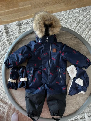 Ny Kuling vinteroverall strl 80 + matchande mössa & vantar - Fin och varm vinteroverall från Kuling i storlek 80 med sött körsbärsmönster. Vatten- och vindtålig med avtagbar pälskant på huvan. Säljs tillsammans med matchande vantar(strl 0-2 år) och vintermössa (strl 48). hann tyvärr bli för liten innan den kom till användning. Endast tvättad med parfymfritt.