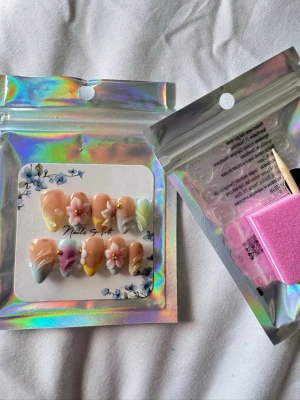 Pastellfärgade press-on naglar med blommor - Säljer ett set press-on naglar från Nails Spot i pastellfärger med 3D-blommor och pärldetaljer. Naglarna har en mjuk mandelform och kommer i nyanser av rosa, gult, lila och mintgrönt. Perfekt för dig som vill ha en söt och kreativ nagelstil.