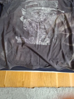 Svart t-shirt med tryck - Cool svart t-shirt med ett stort grafiskt tryck av Tupac på framsidan. T-shirten har korta ärmar och en avslappnad passform. Perfekt för dig som gillar streetwear och vill ha en unik look.