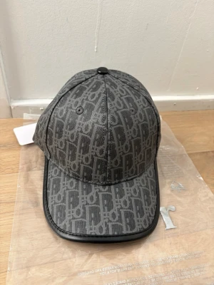 Grå CD keps med monogram - Snygg grå keps från CD med ikoniskt monogrammönster över hela kepsen. Klassisk böjd skärm med svart kant och justerbar baksida. Tillverkad i ett slitstarkt tyg som ger en exklusiv känsla. Perfekt accessoar för dig som gillar streetstyle och lyx. Helt ny, aldrig använt ✅