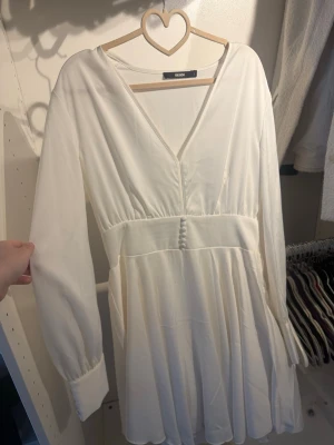 Vit kortklänning från Bik Bok - Supersöt vit kortklänning från Bik Bok med v-ringning, långa ärmar och knappar framtill i midjan. Klänningen har ett flowy fall och markerad midja som ger en feminin siluett. Perfekt för sommarens alla tillfällen!