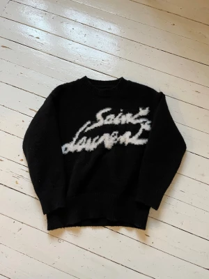 Svart Saint Laurent stickad tröja - Svart stickad tröja från Saint Laurent med vit, fluffig logotyp framtill. Tröjan har rund hals, långa ärmar och ribbstickade muddar vid ärmslut och nederkant. Perfekt för dig som gillar streetstyle och exklusiva märken. Pris kan absolut diskuteras!😃
