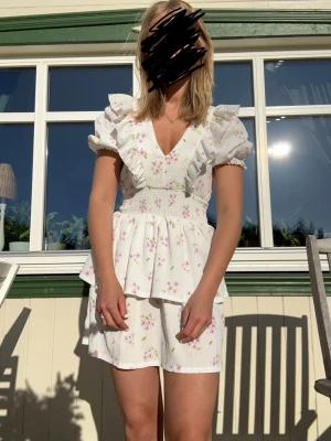 Vit blommig kort klänning med volang - Supersöt vit kort klänning med rosa blommigt mönster, puffiga kortärmar och v-ringning. Från Gina och storlek M.