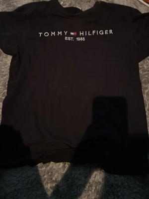 Svart Tommy Hilfiger t-shirt - Klassisk svart t-shirt från Tommy Hilfiger med logga och texten 'EST. 1985' tryckt på bröstet. Enkel och stilren design, perfekt till jeans eller shorts. Tillverkad i mjuk bomull och har korta ärmar.