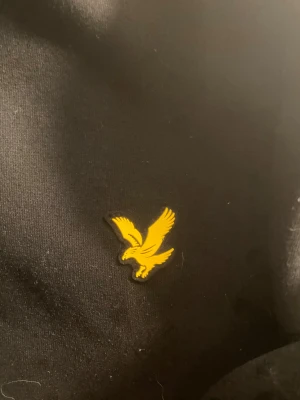 Svart hoodie från Lyle & Scott - Snygg svart hoodie från Lyle & Scott med den klassiska gula fågel-loggan på bröstet. Hoodien har en mjuk känsla och är perfekt för dig som gillar stilrena plagg med en sportig touch. Passar dig som vill ha en enkel men ändå ikonisk look.