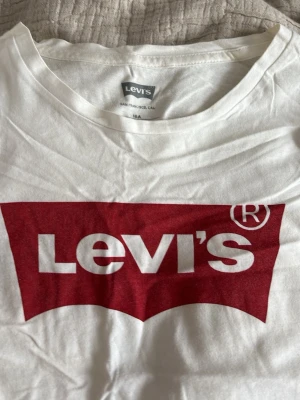 Vit Levi's t-shirt med röd logga - Klassisk vit t-shirt från Levi's med den ikoniska röda loggan tryckt framtill. T-shirten har rund hals och korta ärmar, tillverkad i mjuk bomull för en skön känsla. Perfekt till jeans eller shorts för en avslappnad look.