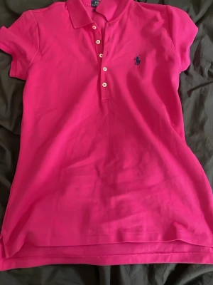 Rosa pikétopp från Ralph Lauren - Klassisk rosa pikétopp från Ralph Lauren med korta ärmar, krage och knappar framtill. Ikoniska blå loggan broderad på bröstet. Tillverkad i mjuk bomull som känns skön mot huden. Perfekt till jeans eller kjol för en sportig och stilren look.