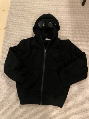 Svart hoodie från C.P. Company - Säljer en svart hoodie från C.P. Company med dragkedja och ikoniska glasögon i huvan. Hoodien har två fickor framtill och ribbade muddar vid ärmar och nederkant. Perfekt för dig som gillar streetwear och vill ha något unikt.