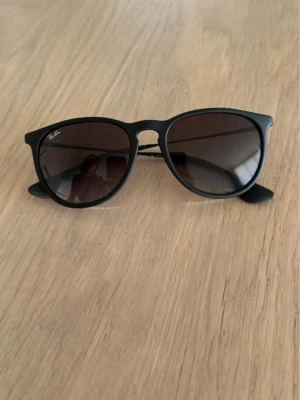 Svarta Ray-Ban solglasögon - Snygga svarta solglasögon från Ray-Ban med klassisk rundad form och mörka glas. Perfekta för att höja din stil och skydda ögonen mot solen. Diskret Ray-Ban-logga i hörnet på ena linsen.