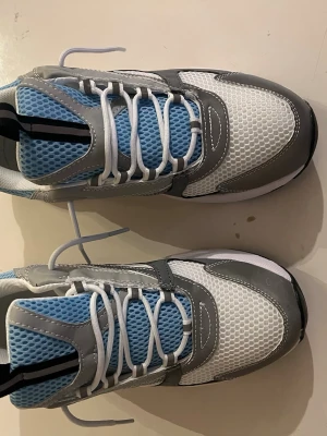 Vita och grå sneakers med blå mesh - Snygga sneakers med vit och grå ovandel i syntetmaterial och mesh. Skorna har blå meshdetaljer på plösen och sidorna, vita snören och svarta detaljer på sulan. Modern design med sportig känsla och bekväm passform.